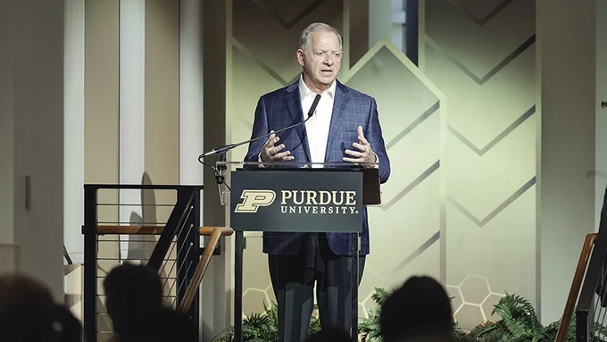 A person speaking at a podium with a Purdue University sign. The background features geometric patterns and plants.
