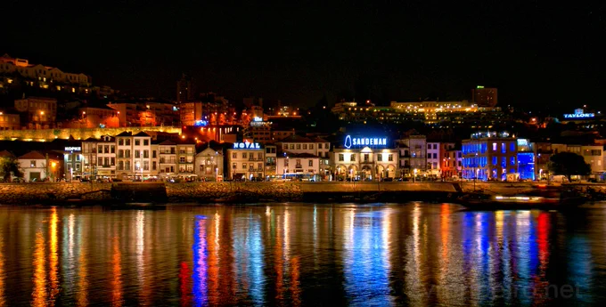 Vista noturna da margem de Gaia com edifícios iluminados e reflexos coloridos no Douro.