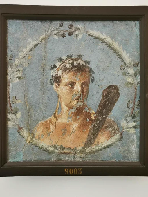FRESCO WITH A CLIPEATE PORTRAIT OF HERAKLES ANTHIOS, 45-79 CE, FROM HERCULANEUM. MUSEO ARCHEOLOGICO NAZIONALE DI NAPOLI.

​Here we have a handsome young Herakles, beardless but identifiable thanks to his clava or club. He is shown within a painted circular wreath against a brilliant blue background. Rather than the traditional laurel, his short, tousled hair is crowned with a delicate wreath of ivy leaves and dark corymbi or berry clusters, a specific iconographical detail that characterises him as Herakles Anthios, the flowered or youthful hero. This version of Herakles was closely linked to festive celebration and the theatre, a fitting patron for a town that claimed the hero as its mythical founder. Discovered during the disordered, unscientific Bourbon-era tunnelings of Herculaneum, the fresco was brutally hacked from its structural context, leaving its exact findspot a mystery. However, the use of exceptionally expensive Egyptian blue pigment strongly points to an elite setting.