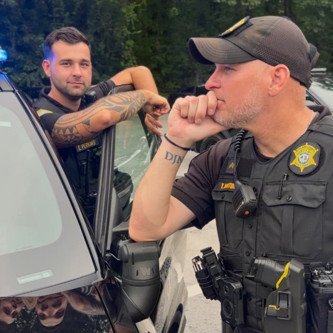 Two uniformed officers standing next to a police vehicle with blue lights on, positioned in a wooded area.
