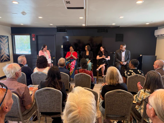 A group of five people sit at the front of a room in a panel discussion setting. They are engaged in conversation, facing an audience seated in rows. Behind the panelists is a large monitor attached to the wall. The room is filled with attendees listening attentively.