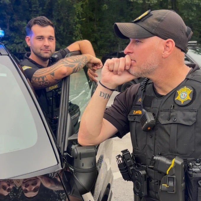 Two uniformed officers standing next to a police vehicle with blue lights on, positioned in a wooded area.