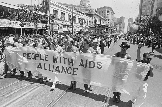 A group of individuals is marching in a parade, holding a large banner that reads "People with AIDS Alliance." A crowd lines the street, and San Francisco's architecture is visible in the background.