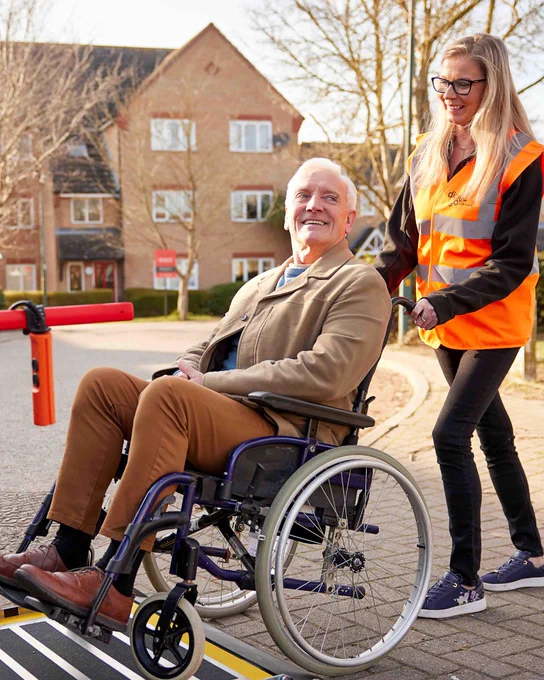A woman pushes an elderly man in a wheelchair on a sunny day, with a residential building in the background.