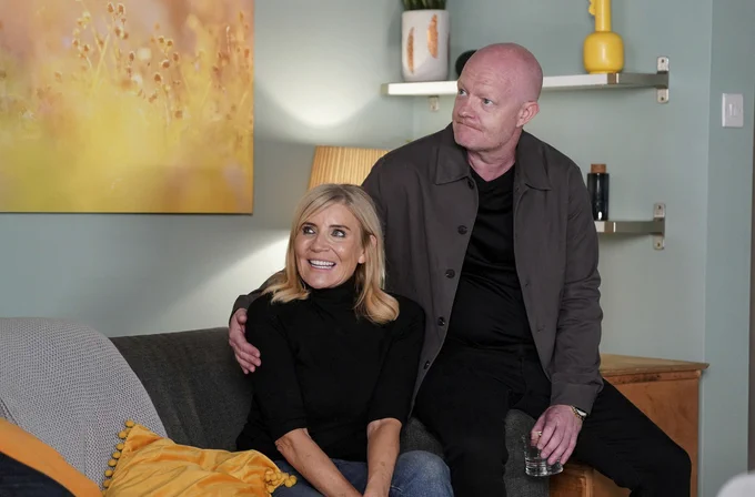 Max sits on the arm of a sofa, placing his arm around Cindy, who is smiling - in a scene from EastEnders.