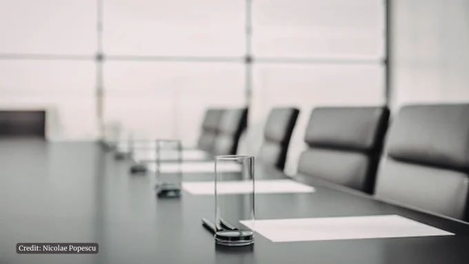 Empty conference room table with neatly arranged glasses and papers, surrounded by black office chairs under soft natural light.