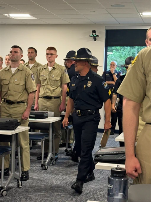 Classroom filled with student officers in tan uniforms and a staff instructor in black addressing the room.