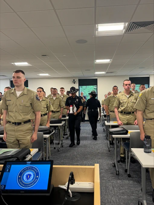 Student officers standing attentively in a classroom with instructors walking down the aisle during training.