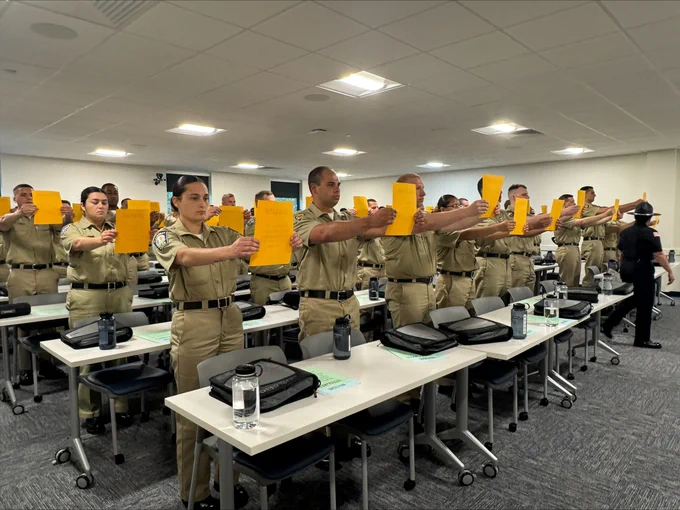 Student officers standing in rows inside a classroom, holding up orange papers during a formal training session.