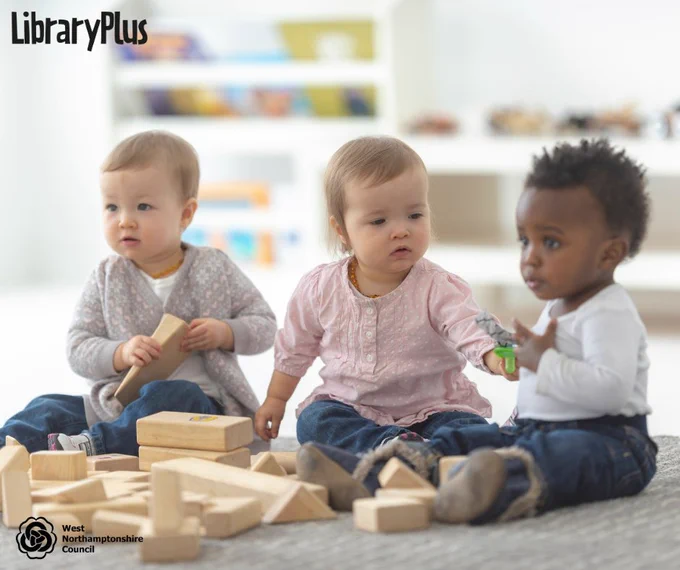 Three toddlers sitting on a carpeted floor playing with wooden building blocks in a bright, child-friendly room.