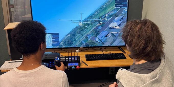 Two LCW students seated at a desk playing a flight simulator game on a large widescreen monitor in a small room.