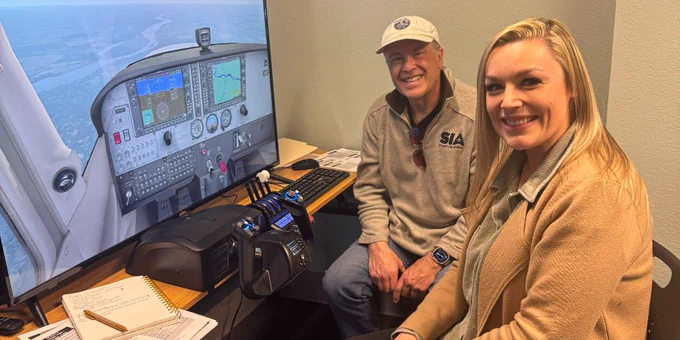 Two people sitting beside a flight simulator setup with a large screen displaying an aircraft cockpit view.