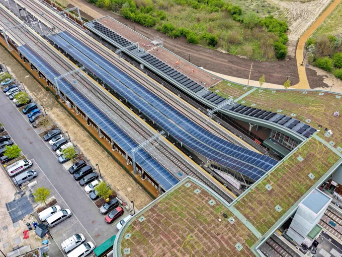 Aerial view of Cambridge South station with platforms covered by solar panels. The station is surrounded by greenery and features a parking lot with several cars. Pathways and grass-covered roofs are visible, enhancing the eco-friendly design.