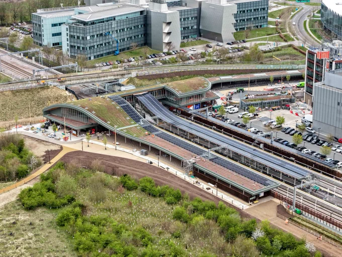 Aerial view of Cambridge South railway station featuring multiple train platforms with overhead shelters. Modern buildings and office complexes are situated nearby, surrounded by roads and green areas.