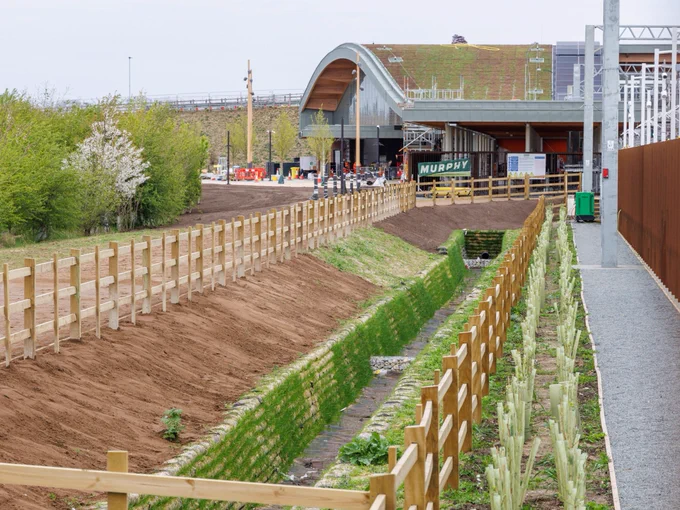 Cambridge South station is seen in the background. In the foreground, there is a landscaped area with young plants and a wooden fence alongside a pathway.