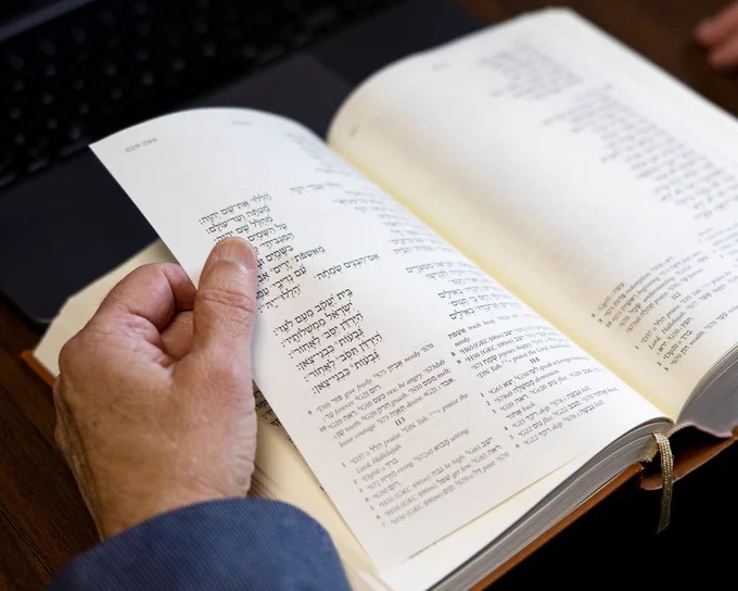 A close-up of a person turning the page of a Hebrew textbook, possibly for study. The setting has a calm and focused atmosphere.