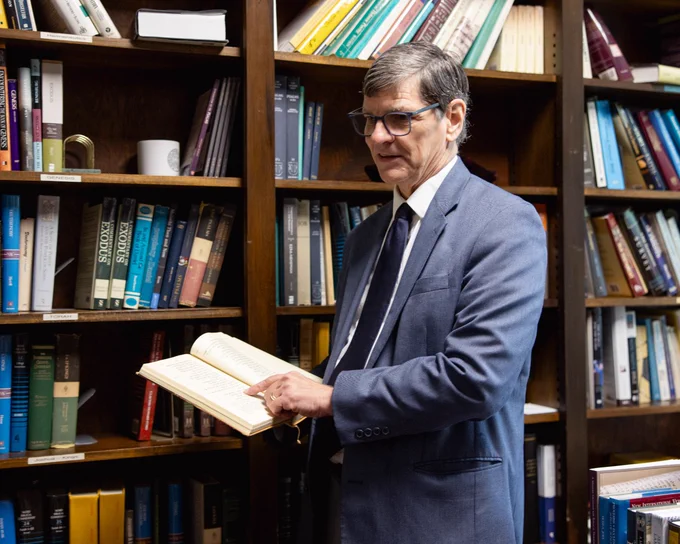 A man in a blue suit is reading a book in a library filled with bookshelves. He looks engaged and focused.