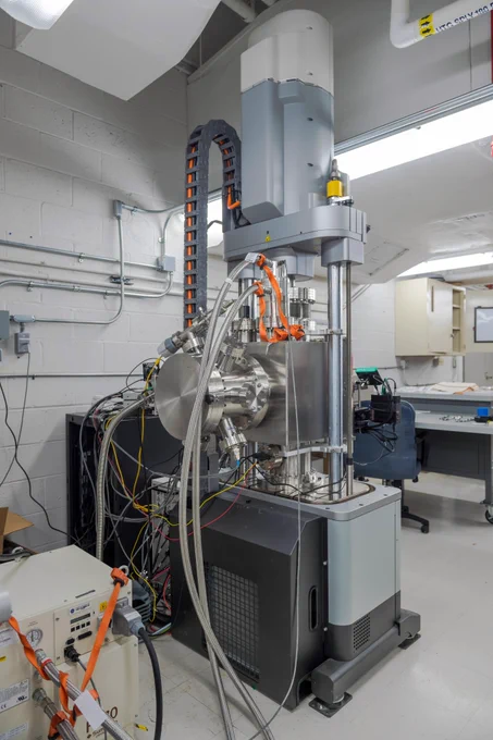 A tall, industrial machine with metallic and gray components stands in a laboratory. Multiple cables and tubes connect to the central chamber, which is cylindrical. Some cables are silver and braided, while others are orange or yellow. The test rig has vertical support columns and a black base. In the background there are white brick walls, ceiling ducts, and a work station with a desk and chair. Credit: NASA/Steven Logan
