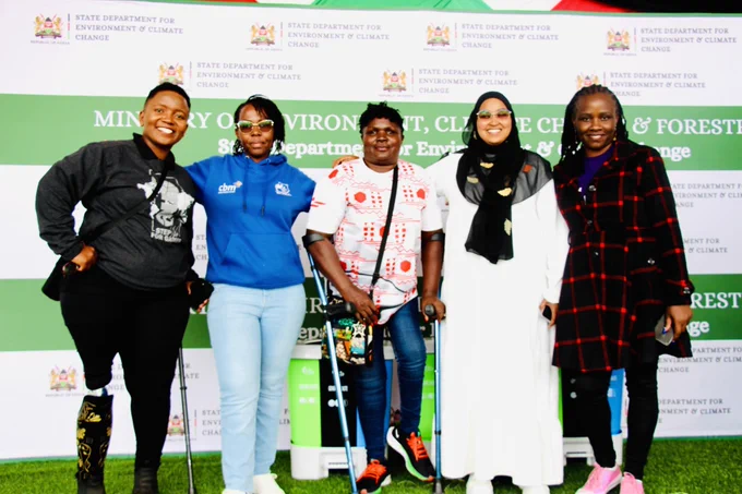 This photo shows a group of five women posing together at  an environment event. They are standing in front of a branded backdrop displaying the logo and name of the State Department for Environment & Climate Change of Kenya.