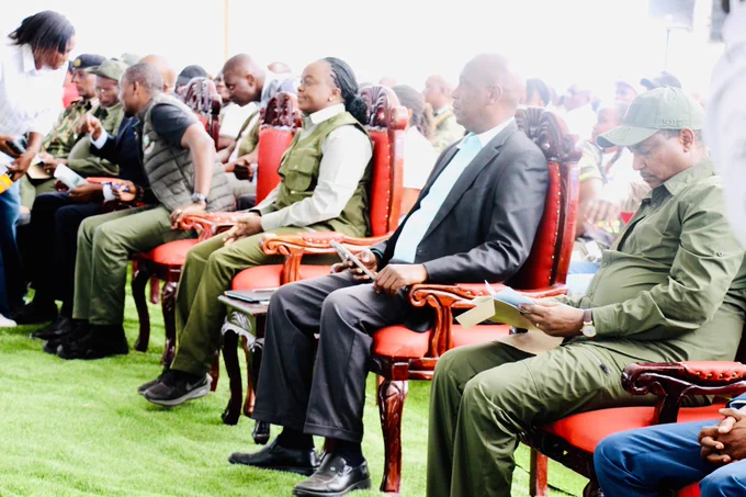 A group of people,some in formal attire and others in military-style uniforms, are seated in red chairs on a grassy field, appearing to be attending an outdoor event.