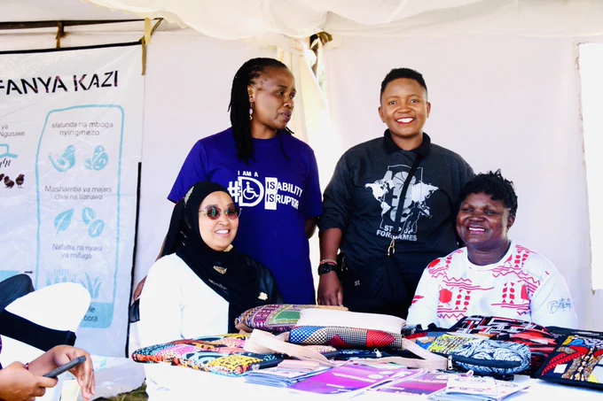 Four individuals are gathered around an exhibition table displaying colorful handmade products, including bags, pouches, and advocacy materials.