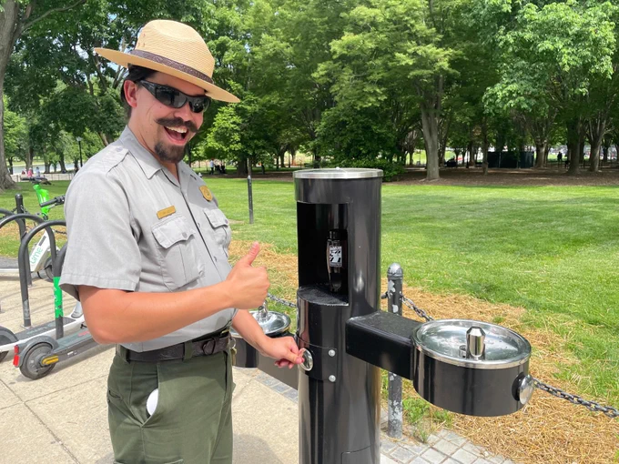 Park ranger in uniform giving thumbs up next to a water fountain with a bottle filler in a park.