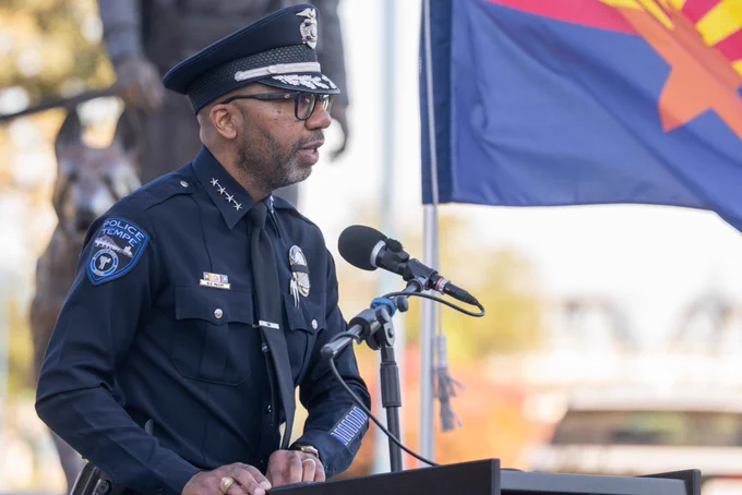 An individual in a police uniform speaks at a podium outdoors. The person is wearing a hat and glasses. Nearby is a microphone, and the Arizona state flag is visible in the background.