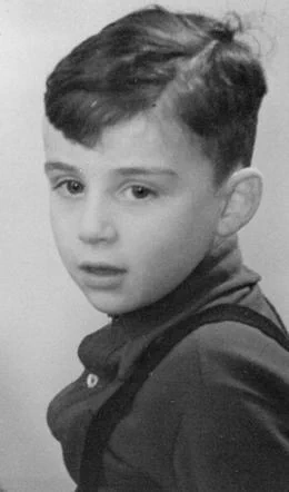 Black and white vintage portrait of a young boy with short hair, looking to the side. The child is wearing a collared shirt underneath a dark sweater. The background is neutral and blurred. 
