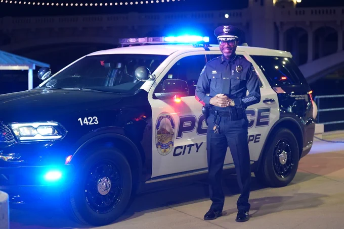 A police officer stands smiling in front of a "City of Tempe" police SUV with flashing lights. The vehicle is parked on a street at night.