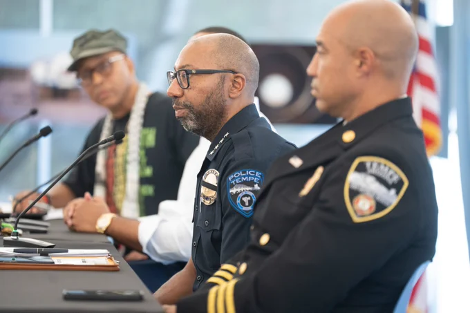 A group of individuals in police uniforms and casual attire are seated at a table with microphones. The setting appears to be a press conference or public meeting with focus on the person in a Tempe Police uniform. The background is blurred, showing an American flag.