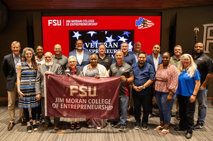 A group of adults posing together holding a maroon 'FSU Jim Moran College of Entrepreneurship' banner in front of a large screen displaying a 'Veteran Entrepreneurship' graphic.