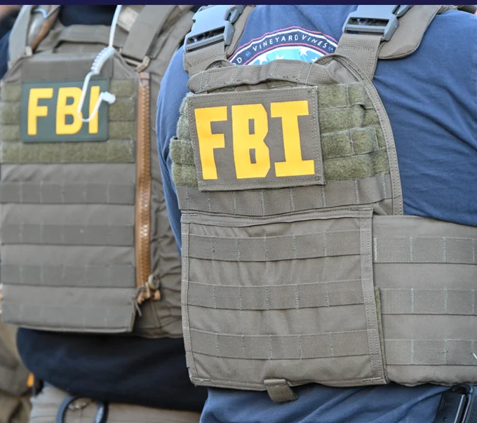 Close-up of two FBI agents wearing tactical vests with bright yellow FBI patches on their backs.