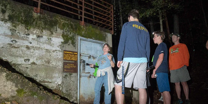 Group of four people standing outside a metal door embedded in a mossy concrete wall at night in a forested area.