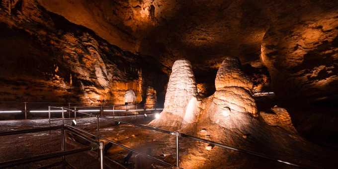 Illuminated stalagmites inside a dimly lit cave with metal railings guiding visitors along the path.