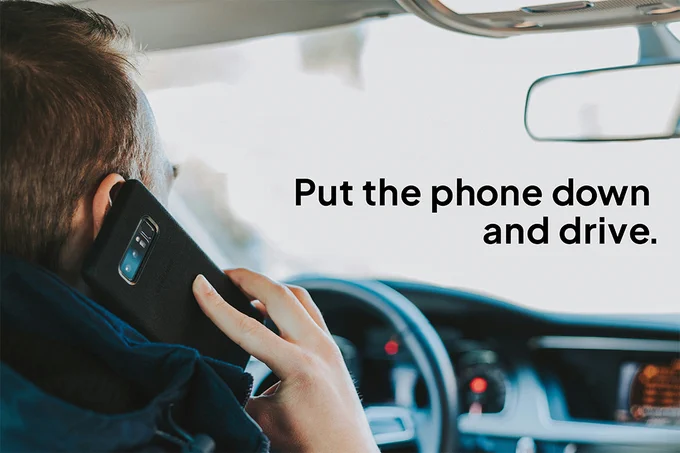 A driver behind the wheel of a vehicle holds a phone to his ear, with the words “Put the phone down and drive.”