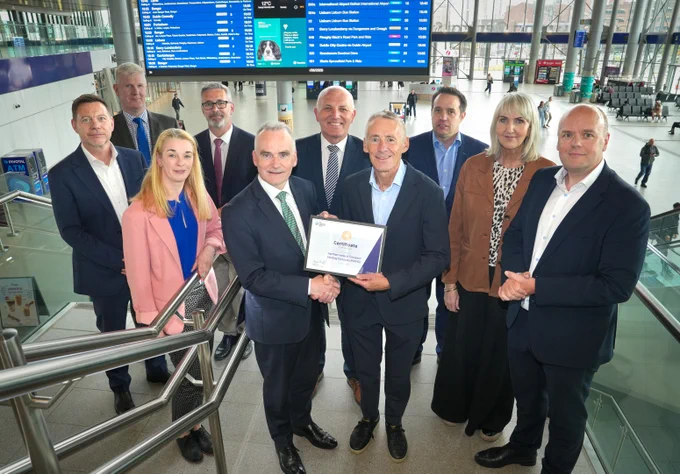 Ten individuals, mostly men in business attire, pose for a photo inside a modern transport hub with glass walls. Two men in the center shake hands, holding a \"Certificate of Membership\" for \"Northern Ireland Transport Holding Company (NITHC)\". Digital screens in the background show transport schedules and weather.