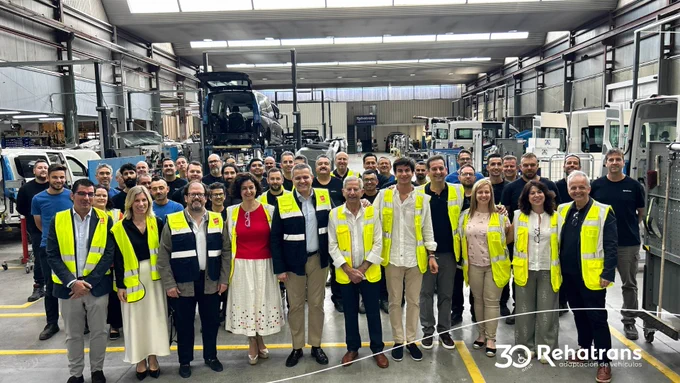 Grupo de trabajadores y personal con chalecos reflectantes posando en un taller de vehículos con furgonetas y elevadores.