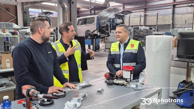 Tres hombres en un taller de adaptación de vehículos, conversando junto a una mesa con piezas y furgonetas elevadas al fondo.