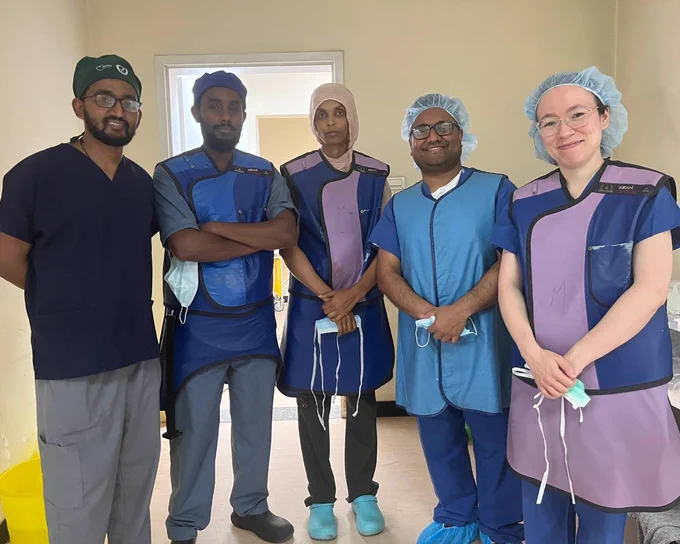 Five people stand together for a picture. They are wearing scrub caps and holding masks in their hands