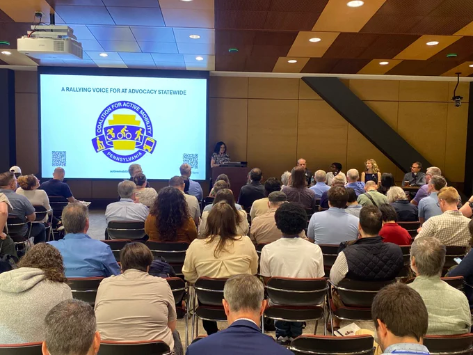 A group of people listening to a presenter speak. There is a PowerPoint behind the presenter that says, " A rallying voice for at advocacy statewide."