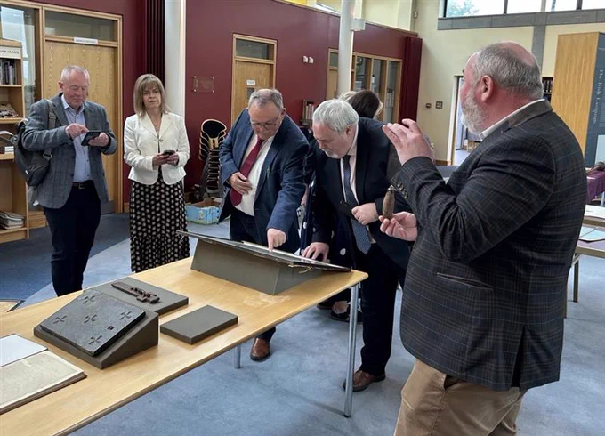 A group of people in Cardinal Tomás Ó Fiaich Library & Archive looking at historic artefacts. The group includes Maolíosa McHugh MLA and Colm Gildernew MLA, Chair of the Committee for Communities.