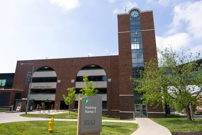 Red brick parking structure with arched openings and a tall glass clock tower on the right. A sign in the foreground reads "Parking Ramp 3" with "OPEN" underneath, and the University of Iowa logo. A tree and fire hydrant are on the grassy area near the entrance. The sky is blue with a few clouds.