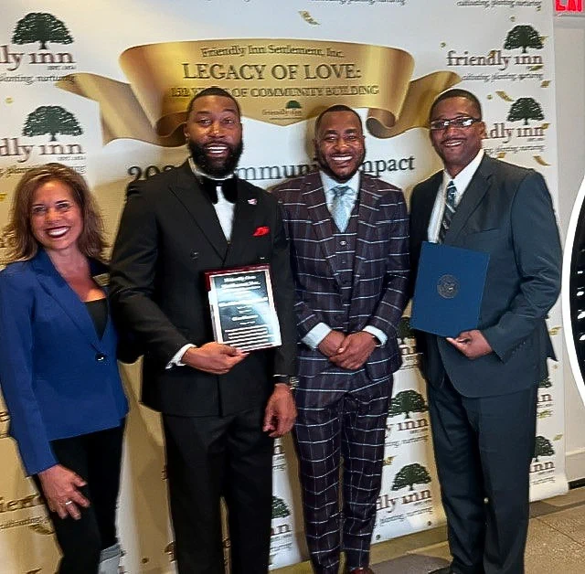 Four people are standing in front of a backdrop that reads "Friendly Inn Settlement, Inc. Legacy of Love: 15 Years of Community Building." One person is holding a plaque, and another is holding a certificate. They are dressed in formal attire and smiling for a group photo.