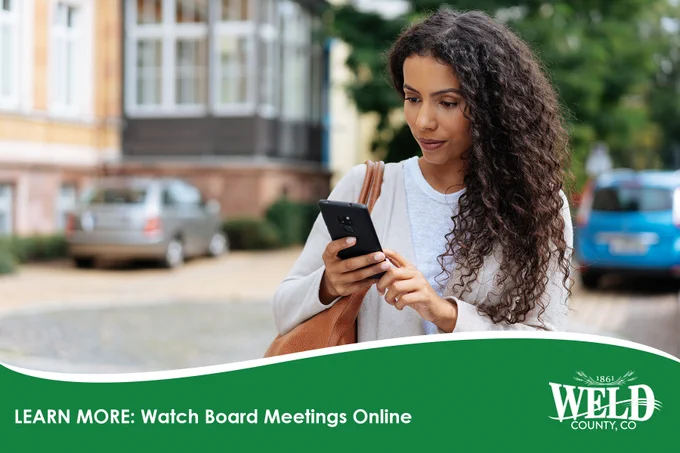 Woman with long curly hair holding a smartphone outdoors near residential buildings.