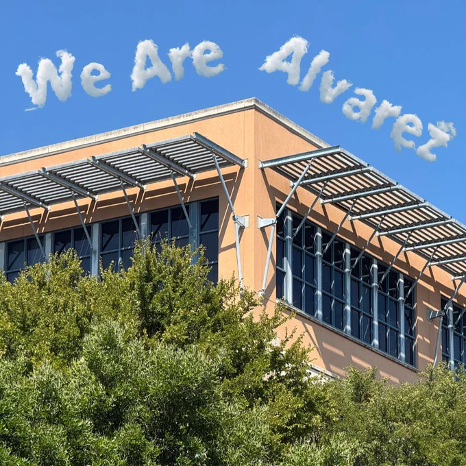 Modern office building with large windows and metal sunshades under a blue sky featuring cloud text saying 'We Are Alvarez'.