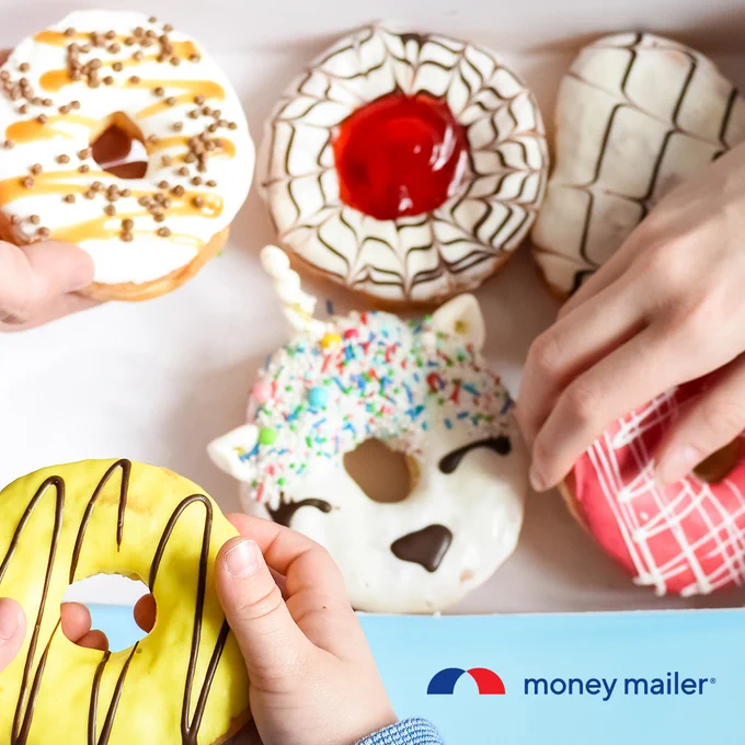 Hands holding colorful donuts with various icing designs, including a unicorn-shaped donut with sprinkles and a red jelly-filled donut on a white surface.