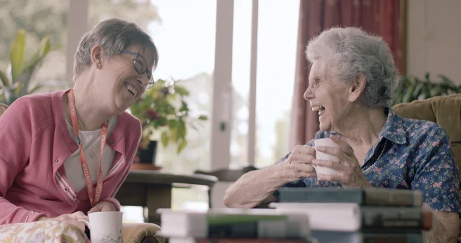 Two women, one a Home Library Direct volunteer, laughing together while having drinks indoors.