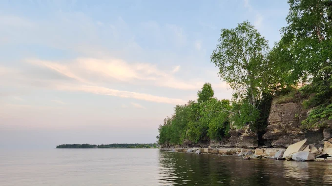 Calm lake beside a rocky cliff shoreline with green trees, still water, and a soft blue sky.