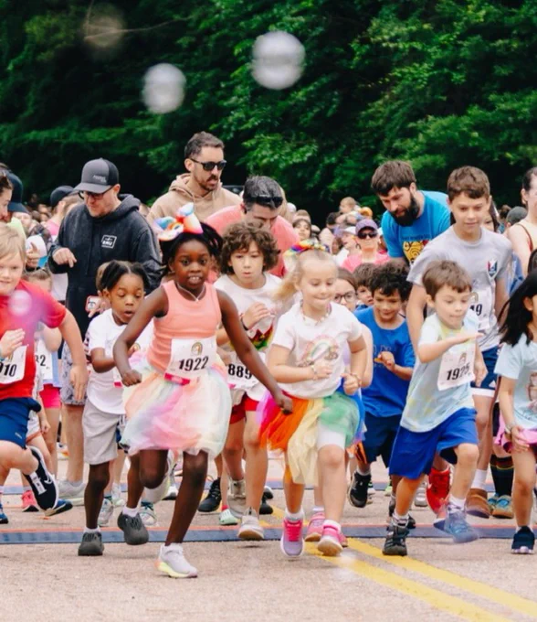 Group of children eagerly starting a race under a large black inflatable finish line arch on a tree-lined road.