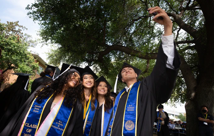 Graduates in their caps and gowns and sashes taking a selfie with trees in the background